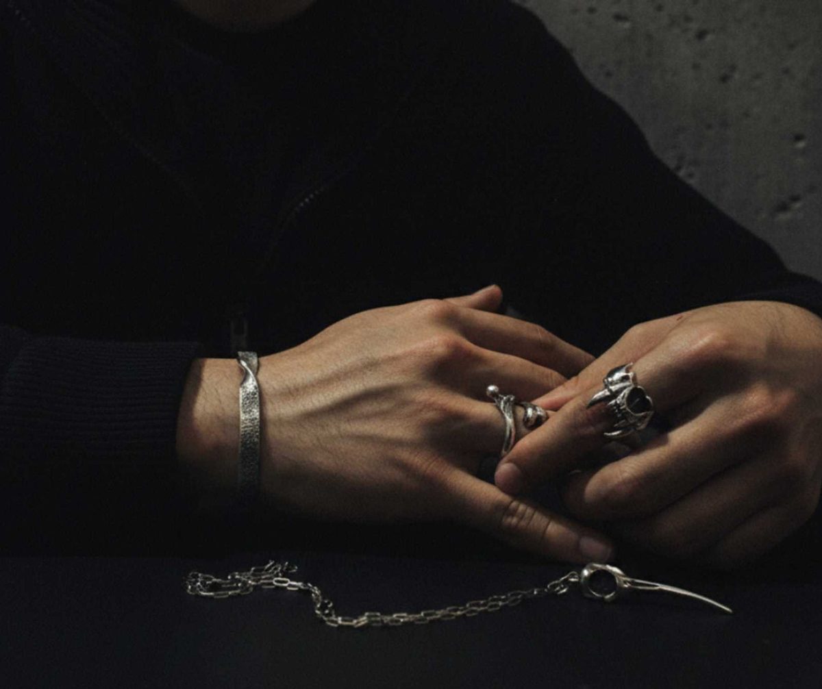 Composición editorial Avalon: Manos con anillos de calavera, brazalete brutalista y collar memento mori sobre mesa de piedra